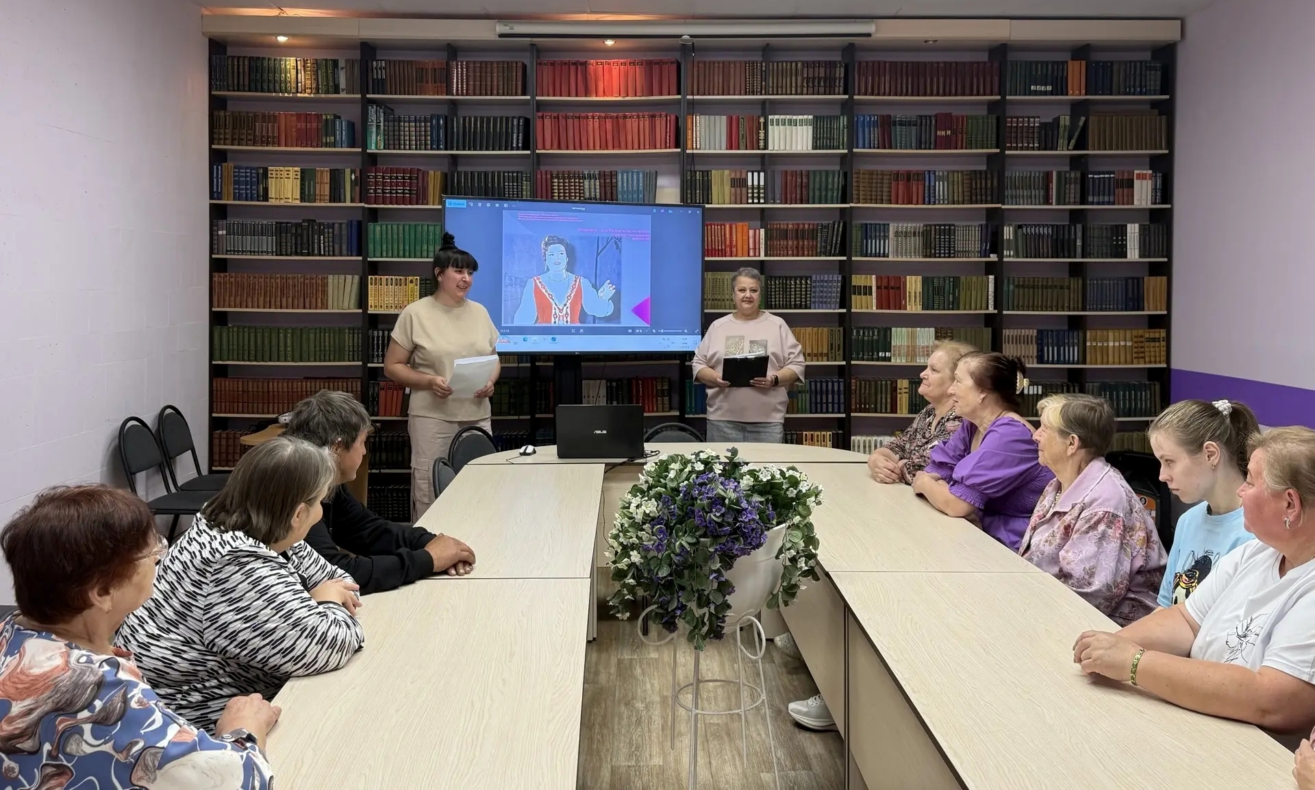 На фото &mdash; встреча в помещении библиотеки. В центре кадра &mdash; длинный светлый стол, за которым сидят слушатели: преимущественно женщины разного возраста, они обращены в сторону выступающих. На столе &mdash; декоративная композиция: в белой вазе на изящной подставке &mdash; букет из искусственных фиолетово‑синих цветов с зелёной листвой.  На фоне &mdash; массивные книжные стеллажи, плотно заполненные книгами в твёрдых переплётах; оттенки обложек варьируются от тёмно‑бордового и коричневого до зелёного и бежевого. Перед стеллажами стоят две женщины‑ведущие: обе в светлой одежде, в руках у них папки или блокноты, они выглядят вовлечёнными в проведение мероприятия. Между ними &mdash; большой экран, на котором транслируется видео: на нём &mdash; ещё одна женщина в этническом наряде. Рядом с экраном на столе стоит ноутбук. Слева у стены &mdash; несколько складных стульев.